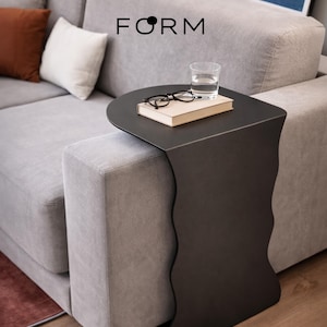 Può includere: Un tavolino nero con un bordo ondulato è posizionato accanto a un divano grigio. Il tavolo contiene un libro, occhiali e un bicchiere d'acqua. La parola "FORM" è stampata in nero sopra il tavolo.