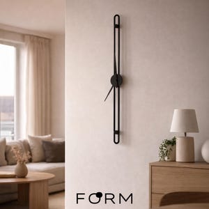 Op de afbeelding: Een moderne zwarte wandklok met een minimalistisch ontwerp. De klok heeft een lang, verticaal frame met een ronde wijzerplaat en wijzers. Het woord "FORM" is in het zwart onderaan de afbeelding gedrukt. De klok is aan een lichtgeleurde muur bevestigd.