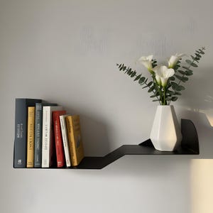 Può includere: Una moderna mensola da parete in metallo nero con un design geometrico unico. La mensola contiene una collezione di libri e un vaso bianco con calle bianche e rami di eucalipto. La mensola è montata su una parete grigio chiaro.