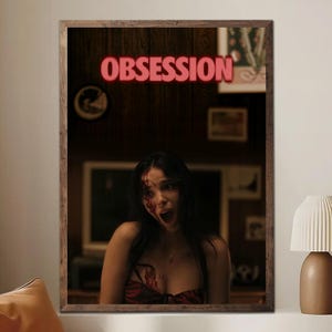 May include: A framed print featuring a woman with blood on her face and chest, screaming with her mouth open. The word "OBSESSION" is in red neon letters above her. The frame is brown, and the background is a dark wood panel.