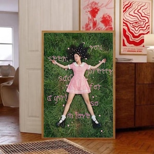 May include: A framed print featuring a person lying on grass, wearing a pink dress and black shoes. The words "seen you pretty sad for a girl so in love" are written in pink. Two other framed prints are visible in the background.