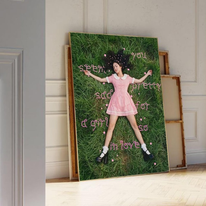 May include: A framed art print featuring a person lying on grass, wearing a pink dress and black shoes. The print has text that reads "seven pretty sad for a girl in love". The artwork is leaning against other canvases.