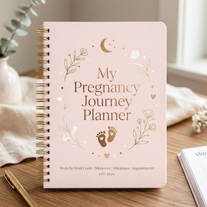 Op de afbeelding: Een roze zwangerschapsplanner met gouden spiraalbinding. De cover toont "My Pregnancy Journey Planner" met bloemen- en maanillustraties. Een open pagina toont "Week 24".