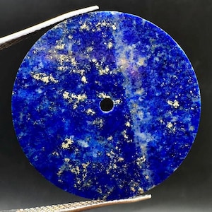Pode incluir: Um pendente circular de lápis-lazúli com uma base de cor azul profunda, veios naturais de calcite branca e inclusões metálicas de pirite dourada. O disco de pedra plano tem um pequeno orifício central para suspensão.