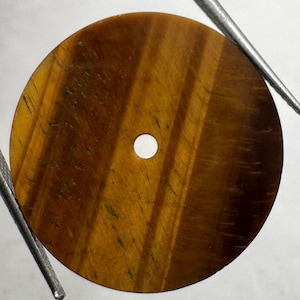 Pode incluir: Uma conta circular de pedra olho de tigre com uma superfície lisa e polida e um orifício central. A pedra apresenta bandas naturais de cor castanho-dourado e castanho-escuro que correm diagonalmente pela sua face plana e redonda.
