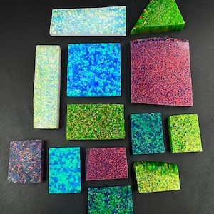 Könnte beinhalten: Eine Sammlung von dreizehn flachen, rechteckigen, quadratischen und dreieckigen synthetischen Opalplatten mit lebendigen, irisierenden Mustern in Blau-, Grün-, Rot- und Violetttönen, die verstreut auf einer dunklen, festen Oberfläche angeordnet sind.