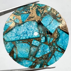 Könnte beinhalten: Ein kreisförmiger Türkis-Edelstein-Cabochon mit einem Mosaikmuster aus hellblauen Steinfragmenten, die von metallisch goldfarbenen Adern durchzogen sind. Das flache, runde Stück hat ein kleines, kreisförmiges Loch nahe der Mitte.
