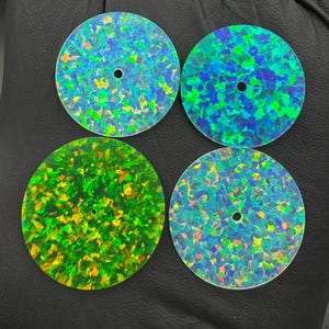 Könnte beinhalten: Vier runde, schillernde Opalsteine mit einem zentralen Loch. Die Steine zeigen ein Mosaik aus Farben, darunter Blau-, Grün- und Gelbtöne. Die Steine sind quadratisch angeordnet, jeder mit einem einzigartigen Farbmuster.
