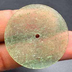 Puede incluir: Un colgante circular de piedra verde translúcida con forma de disco plano y un pequeño orificio circular centrado. La superficie presenta una textura moteada con diversos tonos de inclusiones minerales de color verde claro y oscuro.