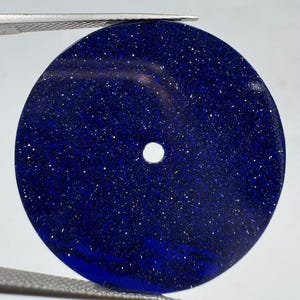May include: A circular, flat blue goldstone disc featuring a deep navy blue base colour with embedded reflective metallic flecks that create a sparkling, star-like appearance. The disc has a small, centred circular hole for mounting purposes.