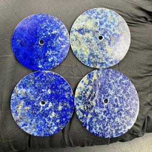 Könnte beinhalten: Vier runde, polierte Lapis-Lazuli-Steine in tiefblauer Farbe mit goldenen Einschlüssen. Jeder Stein hat ein kleines, mittiges Loch. Die Steine sind quadratisch angeordnet.