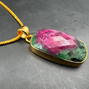 Può includere: Una collana con pendente caratterizzata da una gemma rettangolare sfaccettata di rubino e zoisite verde incastonata in una cornice di metallo dorato. Il pendente è fissato a una catena dorata abbinata con un design a maglie testurizzate.