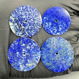 Könnte beinhalten: Vier runde, polierte Lapis-Lazuli-Steine. Jeder Stein ist tiefblau mit goldenen Einschlüssen und einem kleinen Loch in der Mitte. Die Steine liegen auf einer dunklen Oberfläche. Jeder Stein misst ca. 7.6 cm im Durchmesser.