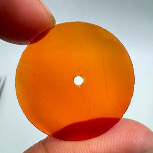 May include: A circular, translucent amber-coloured disc featuring a small, centred hole. The material appears smooth and flat with a consistent orange-brown hue throughout, allowing light to pass through the surface of the round object.