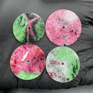 Könnte beinhalten: Vier runde Edelstein-Cabochons mit einem Loch in der Mitte. Die Steine weisen eine Mischung aus Farben auf, darunter Rosa, Grün, Schwarz und Weiß. Die Cabochons sind wahrscheinlich für die Schmuckherstellung oder andere Bastelprojekte bestimmt.