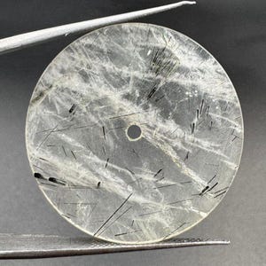 May include: A clear, round gemstone with black inclusions and a central hole. The stone is held by a silver tool, showcasing its polished surface and natural patterns. The gemstone's design is circular.