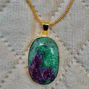 Könnte beinhalten: Eine goldfarbene Halskette mit einem ovalen Anhänger mit polierter Oberfläche. Der Anhänger zeigt einen Naturstein mit ausgeprägten Flecken in tiefem Violett und leuchtendem Grün, gehalten von einer einfachen goldfarbenen Metallöse und Kette.