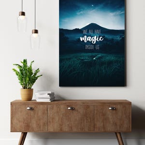 Puede incluir: Un mueble de madera con tres cajones y patas anguladas, coronado por una planta en maceta, libros y una impresión enmarcada. La impresión presenta un paisaje montañoso bajo un cielo estrellado con el texto "WE ALL HAVE MAGIC INSIDE US."