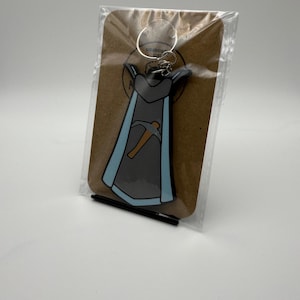 Puede incluir: Un llavero gris y azul con temática de superhéroe con un diseño de pico. El llavero está unido a un llavero plateado y está empaquetado en un respaldo de cartón marrón.