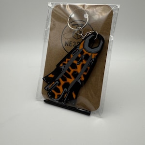 May include: A black and orange keychain with a unique design, attached to a metal ring. The keychain is packaged on a brown card with the text "NE3D" visible. The design features a pattern of orange shapes on a black background.
