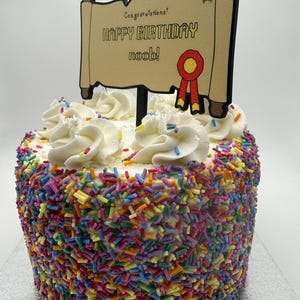 May include: A round birthday cake covered in colorful sprinkles. The cake is topped with white frosting swirls and a decorative sign that reads "Happy Birthday noob!" in black text. A red and yellow ribbon is also on the sign.