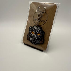 Könnte beinhalten: Ein Schlüsselanhänger mit einem detaillierten Bild eines Helms mit leuchtend orangefarbenen Augen. Der Schlüsselanhänger ist an einem Metallring und einer Kette befestigt und wird auf einer braunen Karte mit dem Text "NEED CO" präsentiert.