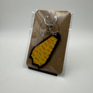 Puede incluir: Un llavero pixelado en forma de mazorca de maíz con detalles naranjas y amarillos, sujeto a un llavero plateado. El llavero está empaquetado en una tarjeta marrón con el texto "NE3D" impreso.