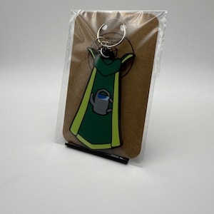 Pode incluir: Um chaveiro de capa de super-herói verde e amarelo com um porta-chaves prateado. O design da capa inclui um regador cinzento. O chaveiro é embalado num cartão castanho numa manga de plástico transparente.