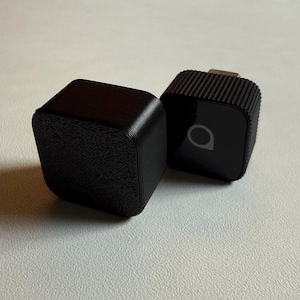 Könnte beinhalten: Zwei schwarze, würfelförmige elektronische Geräte. Eines hat eine strukturierte Oberfläche, das andere hat eine geriffelte Oberseite und ein kleines Logo. Beide befinden sich auf einer hellen Oberfläche. Die Geräte scheinen kleine elektronische Zubehörteile zu sein.