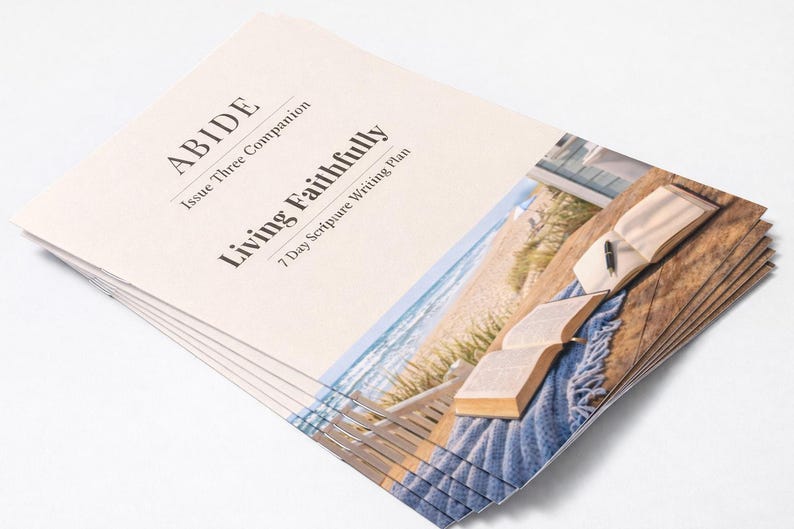 Peut inclure: Une pile de livrets imprim&eacute;s avec le titre "ABIDE" et "Living Faithfully". La couverture pr&eacute;sente une image d'un livre ouvert, d'un stylo et d'une sc&egrave;ne de plage. Les livrets sont empil&eacute;s sur une surface blanche.