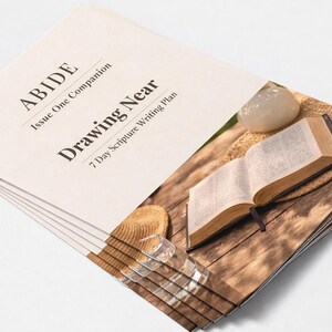 Peut inclure: Une pile de livrets cr&egrave;me avec le titre "ABIDE Drawing Near" et le sous-titre "7 Day Scripture Writing Plan". Un livre ouvert avec un signet, un petit chapeau et un verre transparent sont sur une surface en bois.