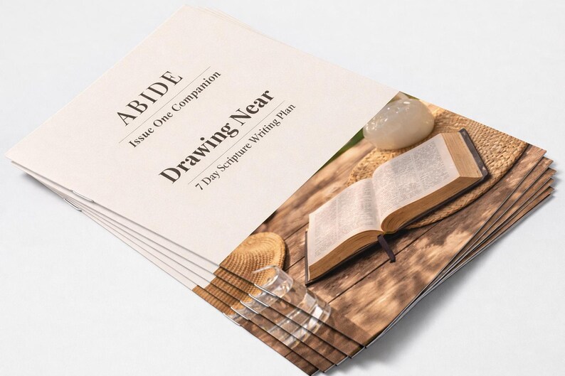 Peut inclure: Une pile de livrets cr&egrave;me avec le titre "ABIDE Drawing Near" et le sous-titre "7 Day Scripture Writing Plan". Un livre ouvert avec un signet, un petit chapeau et un verre transparent sont sur une surface en bois.