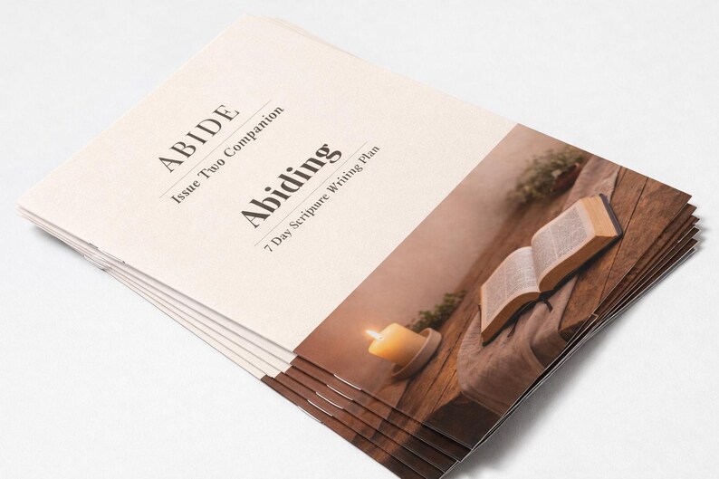 Peut inclure: Une pile de livrets cr&egrave;me avec le titre "ABIDE" et "Abiding" imprim&eacute;s en noir. La couverture pr&eacute;sente une image d'un livre ouvert, d'une bougie allum&eacute;e et de verdure sur une surface en bois. Les livrets sont pour un plan d'&eacute;criture de 7 jours.