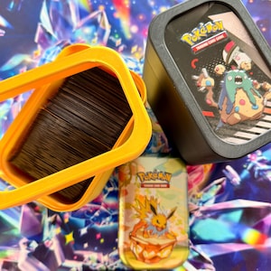 Op de afbeelding: Een feloranje plastic kaartenopbergdoos met een stapel donkere ruilkaarten, naast een zwarte rechthoekige houder met het Pokemon Trading Card Game logo en een Garbodor-illustratie, en een kleine gele Pokemon-blikken doos.