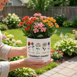 May include: A white ceramic flower pot with a colorful arrangement of red, pink, and orange flowers. The pot is decorated with floral illustrations and the text "Grandma's Garden" and "Love grows here".