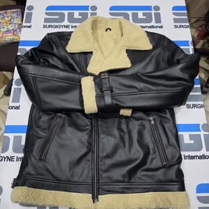 May include: A black jacket with a beige faux shearling collar, cuffs, and hem. The jacket features a front zipper closure, two zippered side pockets, and a buckled strap detail on the cuffs. The design includes a wide fold-over collar.