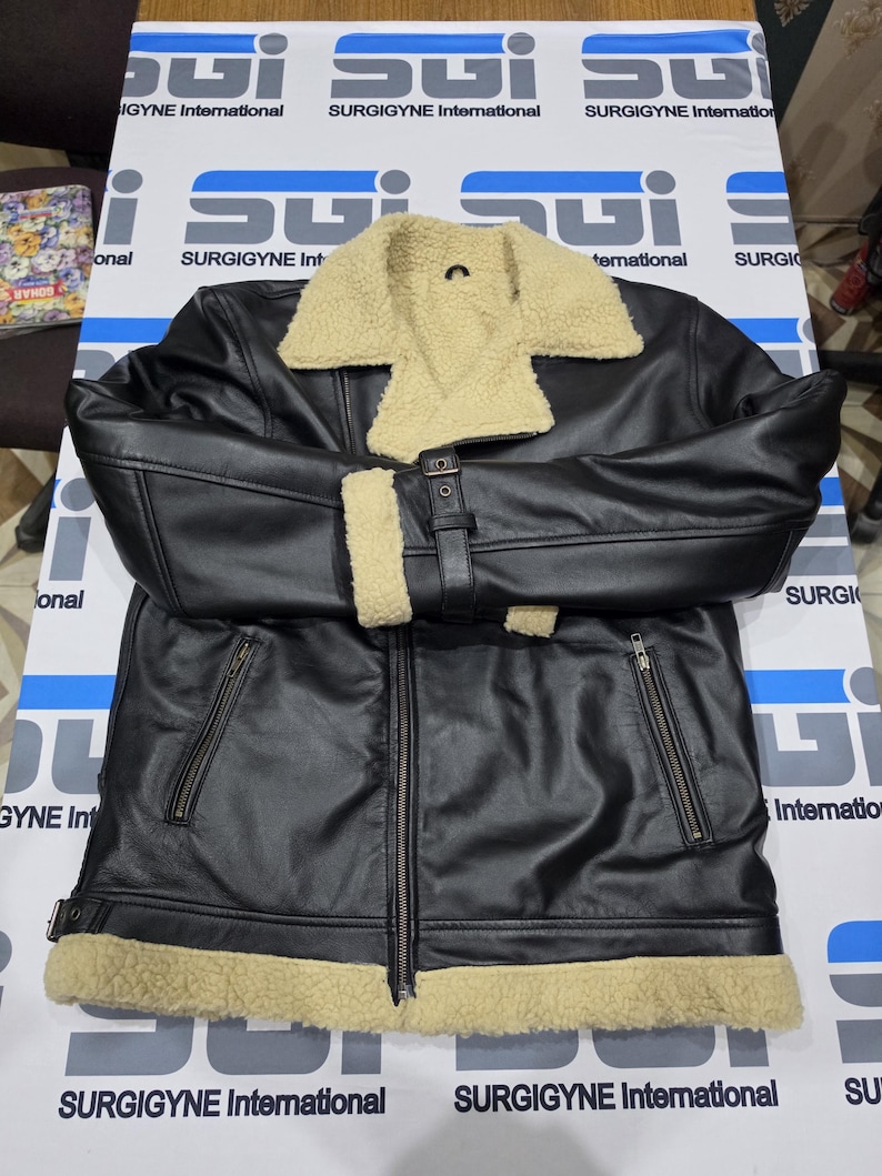 May include: A black jacket with a beige faux shearling collar, cuffs, and hem. The jacket features a front zipper closure, two zippered side pockets, and a buckled strap detail on the cuffs. The design includes a wide fold-over collar.
