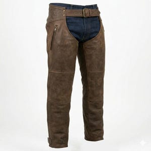 May include: Brown leather chaps with a belt and buckle. The chaps feature a zip pocket on the left leg and are worn over blue denim jeans. The leather has a distressed appearance.
