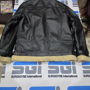 May include: A black leather jacket featuring a beige shearling collar and matching shearling trim along the bottom hem and sleeve cuffs. The jacket includes adjustable buckle straps at the waist and cuffs, with visible seam detailing across the back.