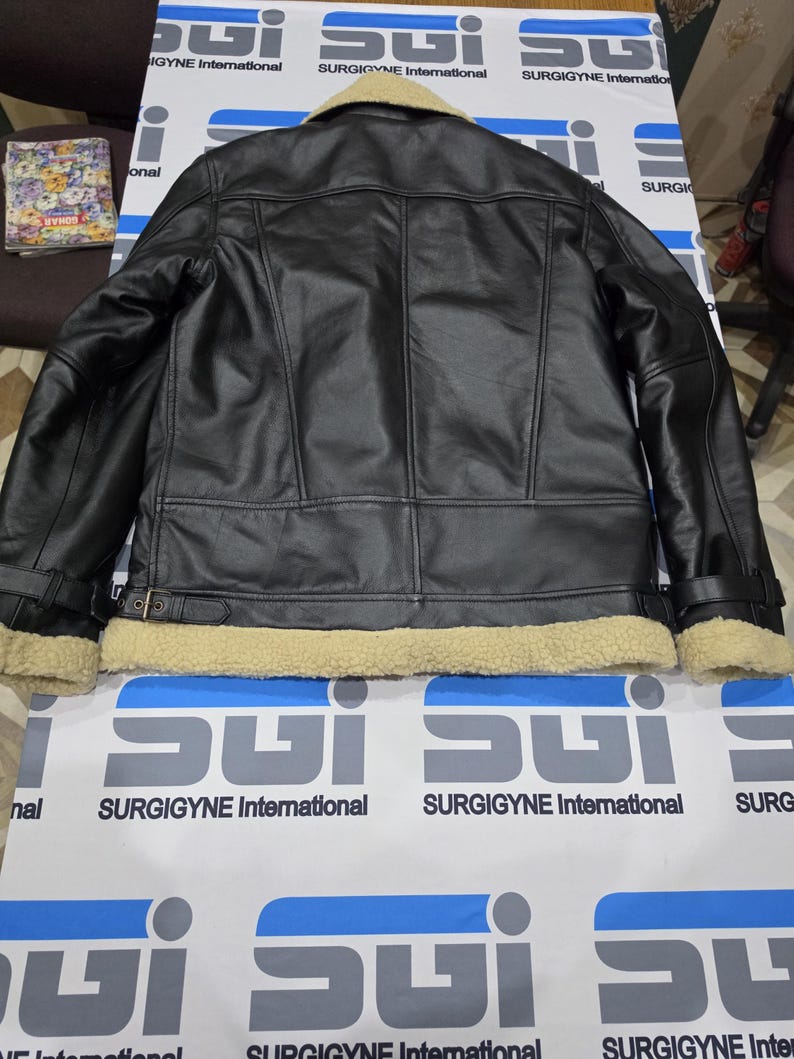 May include: A black leather jacket featuring a beige shearling collar and matching shearling trim along the bottom hem and sleeve cuffs. The jacket includes adjustable buckle straps at the waist and cuffs, with visible seam detailing across the back.