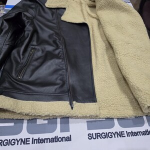 May include: A black leather jacket featuring a beige sherpa lining, a wide fold-over collar, and a front zipper closure. The jacket includes a zippered pocket on the front and is lined with a thick, textured cream-colored fleece material.