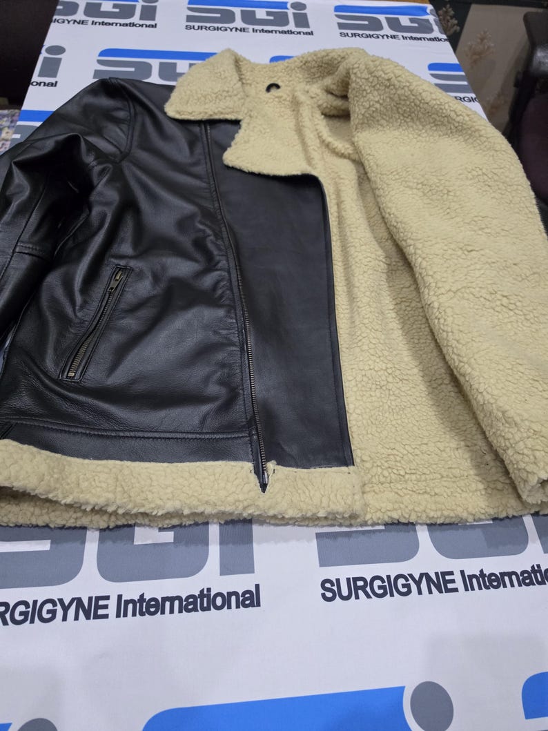 May include: A black leather jacket featuring a beige sherpa lining, a wide fold-over collar, and a front zipper closure. The jacket includes a zippered pocket on the front and is lined with a thick, textured cream-colored fleece material.