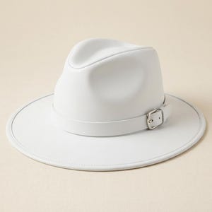 May include: A solid white fedora hat featuring a wide brim and a matching white hat band. The band is secured with a silver-toned metal buckle on the side. The hat has a structured crown with a central crease and a smooth, matte finish.