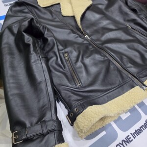 May include: A black leather jacket featuring a cream-colored faux shearling collar and cuffs. The jacket includes a front metal zipper closure, two zippered side pockets, and adjustable buckled straps at the waist and sleeve cuffs.