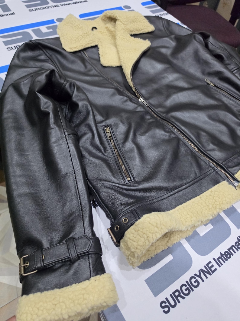 May include: A black leather jacket featuring a cream-colored faux shearling collar and cuffs. The jacket includes a front metal zipper closure, two zippered side pockets, and adjustable buckled straps at the waist and sleeve cuffs.