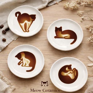 May include: Four white ceramic coasters with brown cat designs. Each coaster features a different cat pose, with the brand name "Meow Ceramics" printed at the bottom. The coasters are arranged on a light wooden surface.