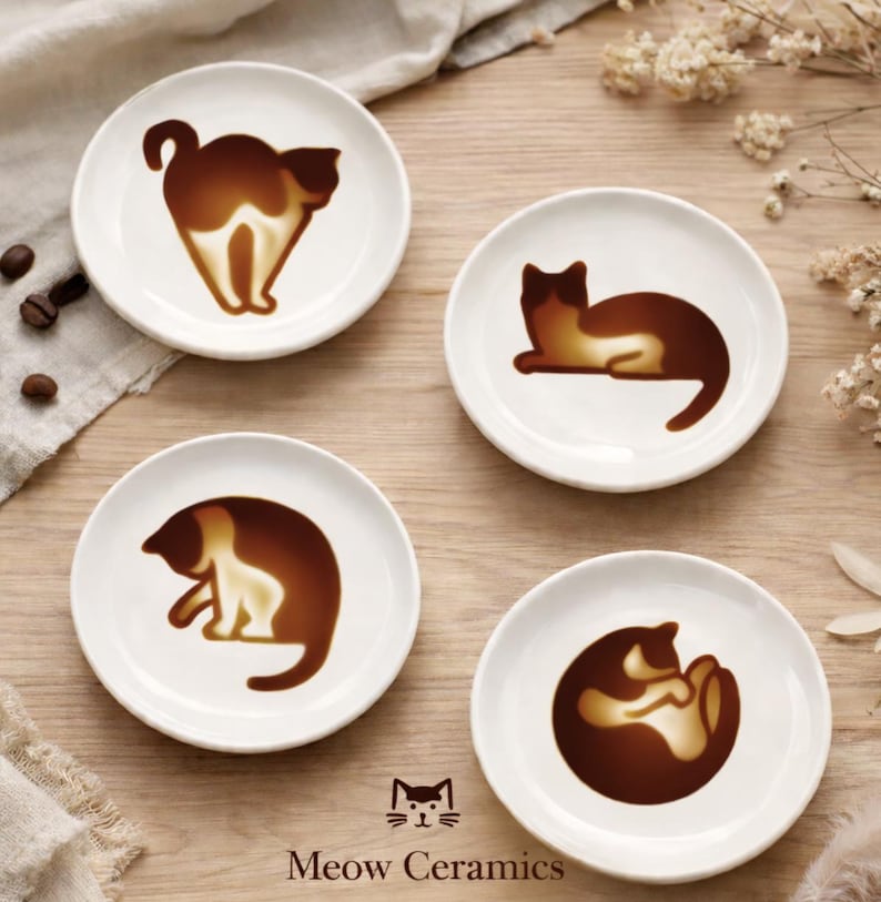 K&ouml;nnte beinhalten: Vier wei&szlig;e Keramikuntersetzer mit braunen Katzenmotiven. Jeder Untersetzer zeigt eine andere Katzenpose, mit dem Markennamen "Meow Ceramics" unten aufgedruckt. Die Untersetzer sind auf einer hellen Holzoberfl&auml;che angeordnet.