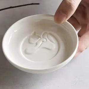 K&ouml;nnte beinhalten: Eine kleine, wei&szlig;e Keramikschale mit einem gepr&auml;gten Pferdekopf-Design in der Mitte. Die Schale ist rund mit einem leicht ausgestellten Rand. Die Schale wird von einer Hand gehalten.