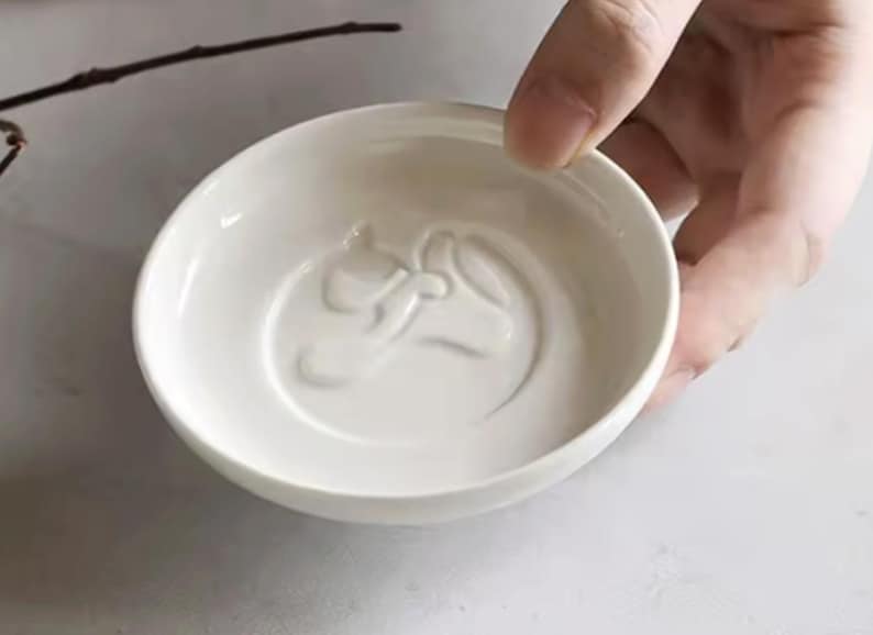 K&ouml;nnte beinhalten: Eine kleine, wei&szlig;e Keramikschale mit einem gepr&auml;gten Pferdekopf-Design in der Mitte. Die Schale ist rund mit einem leicht ausgestellten Rand. Die Schale wird von einer Hand gehalten.
