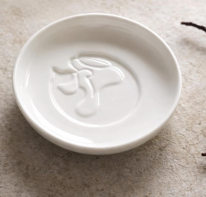 K&ouml;nnte beinhalten: Eine kleine, runde, wei&szlig;e Keramikschale mit einem erhabenen, abstrakten Blumendesign in der Mitte. Die Schale hat eine glatte, gl&auml;nzende Oberfl&auml;che und ist vor einem neutralen Hintergrund platziert. Das Design ist eine stilisierte Blume.